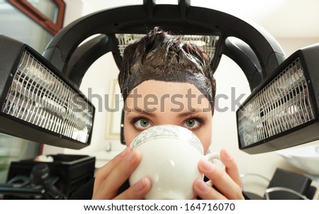 Young woman drinking coffee tea in hairdressing beauty salon. Girl dying colouring hair by hairdresser hairstylist. Modern equipment.