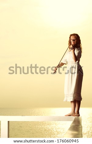 The blonde girl music lover on pier with a violin at sunset or sunrise.  Love of music concept.