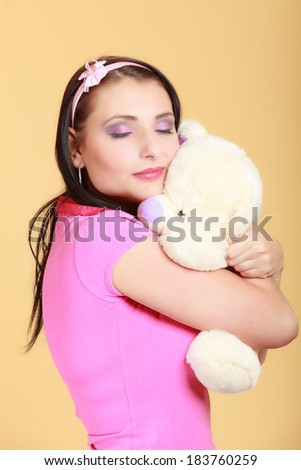 Portrait of childish young woman with headband holding toy. Infantile girl in pink hugging teddy bear on orange. Longing for childhood. Studio shot.