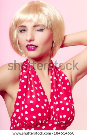 Vintage pinup style. Portrait of beautiful stylized young woman winking. Attractive girl in blond wig and retro spotted red dress on pink. Disguise. Studio shot.