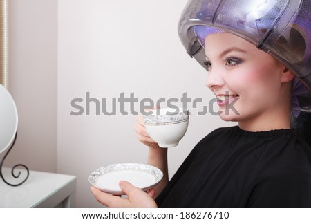Young woman female client drinking hot drink coffee tea in hairdressing beauty salon. Girl in hair rollers curlers with hairdryer dryer relaxing by hairdresser hairstylist.