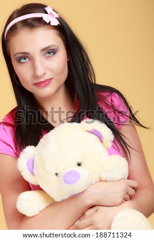 Portrait of childish young woman with headband holding toy. Infantile girl in pink hugging teddy bear on orange. Longing for childhood. Studio shot.