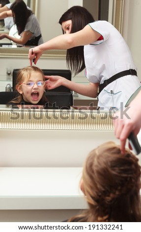 Kid by hairstylist. Hairdresser with comb combing hair of little girl in eyeglasses. Child in hairdressing beauty salon.