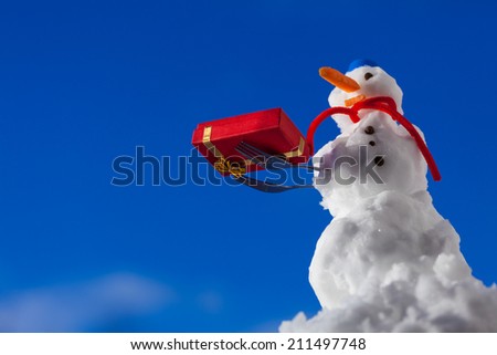 Little happy snowman in blue screw top as hat red scarf and with fork christmas gift box outdoor. Winter season seasonal specific. Blue sky background.