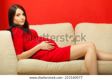 Pregnancy, motherhood and happiness concept. Beautiful sexy stylish elegant pregnant woman in red dress relaxing on sofa and touching her belly