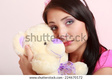 Portrait of childish young woman with headband holding toy. Infantile girl hugging teddy bear on pink. Longing for childhood. Studio shot.