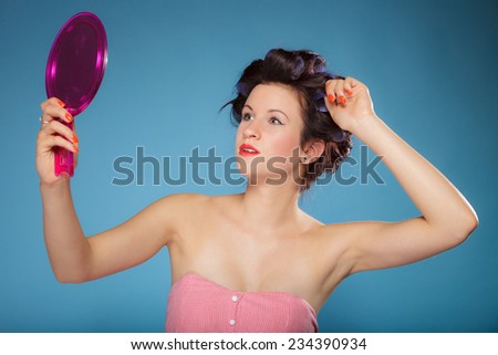 young woman preparing to party having fun, girl styling hair with curlers looking in the mirror retro style blue background
