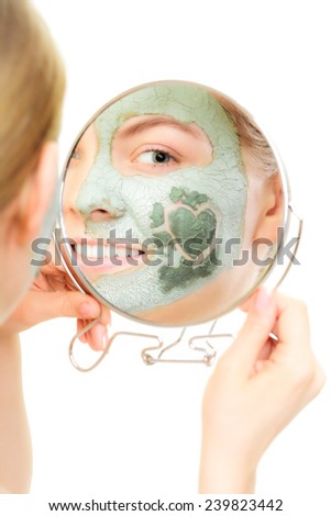 Skin care. Woman in clay mud mask on face with heart on cheek looking in the mirror isolated on white. Girl taking care of dry complexion. Beauty treatment.