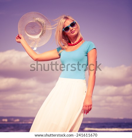 Fashion, happiness and lifestyle concept. Lovely blonde girl in hat and blue sunglasses red beads walking on beach. Young woman relaxing on the sea coast.