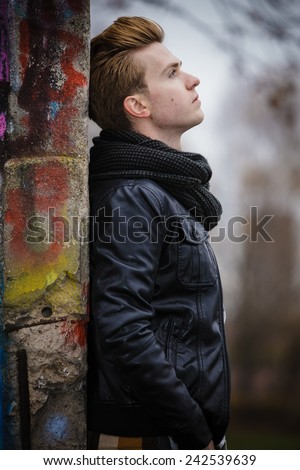 Face profile thoughtful trendy man outdoor in city setting, male model wearing winter clothes black jacket and scarf against colorful graffiti wall
