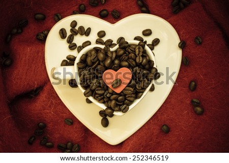Coffee klatsch java concept. Heart shaped white cup filled with roasted coffee beans on red cloth background. Vintage filter