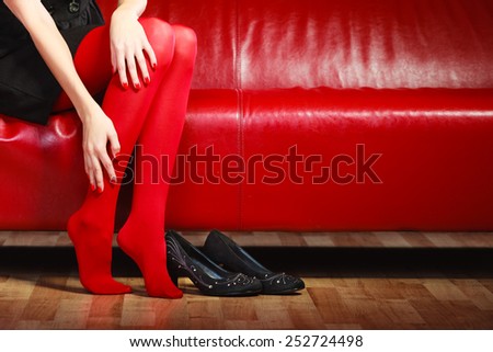 Elegant fashion outfit. Fashionable woman long legs in red vivid color pantyhose black shoes sitting on couch indoor