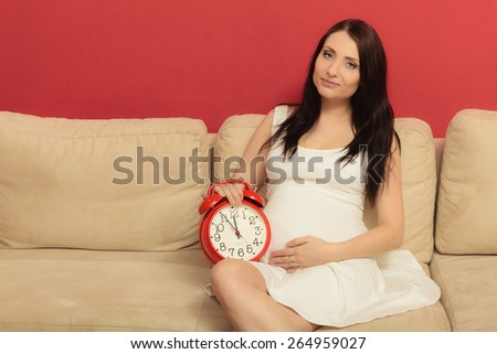 Pregnant woman holding red alarm clock waiting, sitting on sofa at home