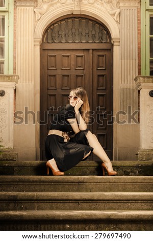 Attractive woman sitting on steps outdoors. Retro style fashionable girl in black classic dress and sunglasses relaxing on street of the old town city Gdansk Danzig Poland