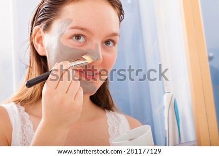 Beauty procedures skin care concept. Young woman no makeup applying facial gray mud clay mask to her face in bathroom using brush