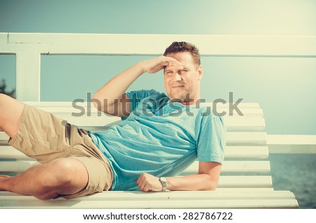 Handsome man tourist laying on bench on pier. Guy enjoying summer travel vacation by sea. Fashion and relaxation.