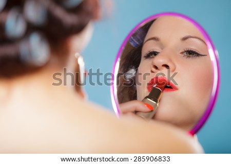 young woman preparing to party, getting ready for going out. Girl styling hair with curlers applying make up red lipstick looking at mirror retro style blue background