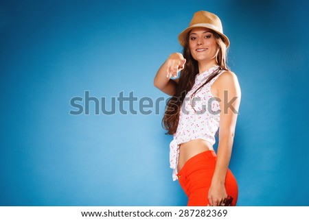 Youth and vacation concept. Happy teen girl in summer clothes and straw hatpointing your with finger. Portrait of smiling beauty woman tourist on blue.