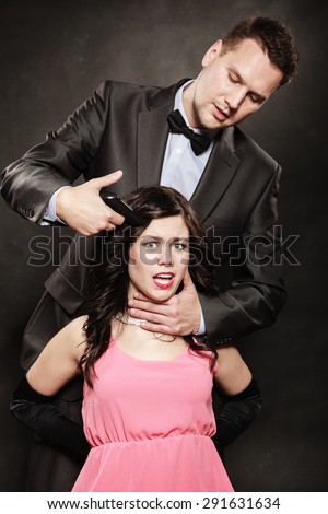 Scene of violence with firearm between men and women. Elegant man holding gun against sitting lady on black and grey background in studio.