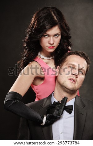 Scene of violence with firearm between men and women. Elegant lady holding gun against sitting man in suit on black grey background in studio.