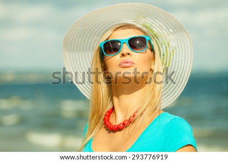 Fashion, happiness and lifestyle concept. Lovely blonde girl in hat and blue sunglasses red beads walking on beach. Young woman relaxing on the sea coast.