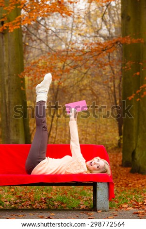 Fall lifestyle concept. Happy crazy blonde woman teen girl having fun in autumn park lying on bench with book.