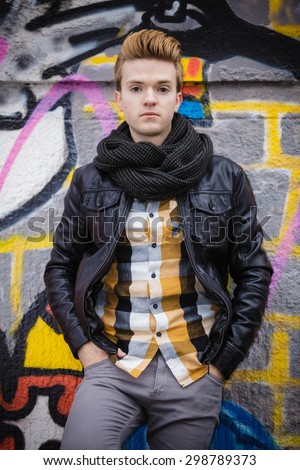 Handsome trendy man outdoor in city setting, male model wearing black jacket scarf and checked shirt against colorful graffiti wall