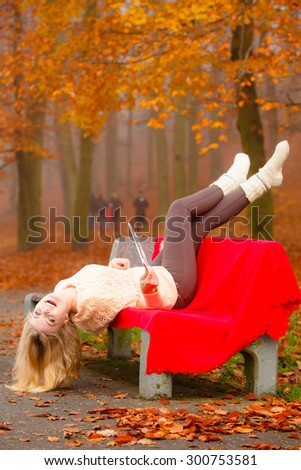 Fall lifestyle concept. Happy crazy blonde woman teen girl having fun in autumn park lying on bench with tablet  e-book reader touchpad pc outdoor