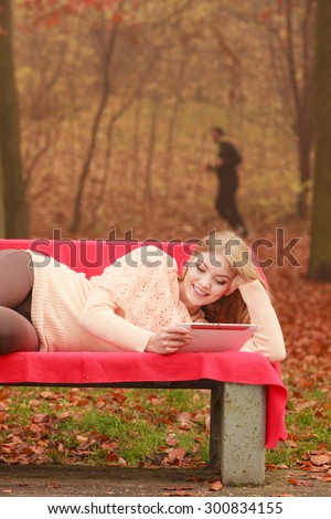 Happy young woman in autumn fall park using computer tablet browsing internet. Girl laying on bench holding pc ebook. Technology leisure.