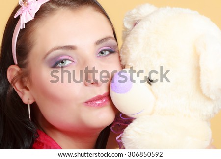 Portrait of childish young woman with headband holding toy. Infantile girl in pink hugging kissing teddy bear on orange. Longing for childhood. Studio shot.