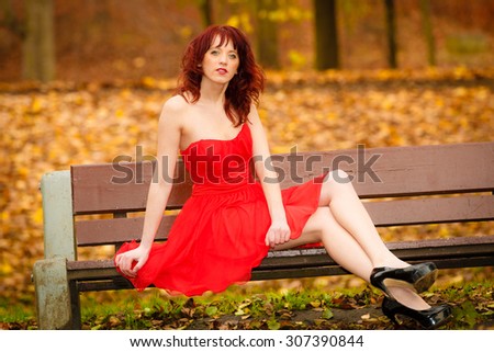 Beautiful young woman in elegant red dress sitting on bench. Girl relaxing on autumnal park. Fall leisure. Outside.