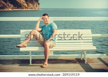 Handsome man tourist sitting on bench on pier. Guy enjoying summer travel vacation by sea. Fashion and relaxation.