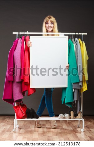 Happy smiling pretty woman in wardrobe holding blank empty banner. Gorgeous girl customer in mall shop with copyspace. Fashion clothing sale advertisement concept.