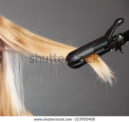 Hairstyling. Closeup blonde woman long haired making hairstyle hairdo with electric hair iron straightener gray background