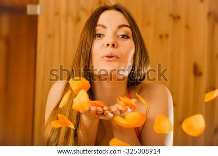 Carefree young woman blowing petals in wood finnish sauna. Attractive girl in bikini relaxing resting. Spa wellbeing pleasure.