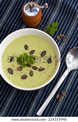 Bowl of broccoli cream soup photographed from top, on a black striped table cloth. Soup is sprinkled with pumpkin seeds and fresh parsley.