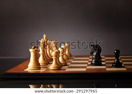 Brave black chess knight and pawn facing the entire army of white chess pieces on a wooden chessboard with dark background