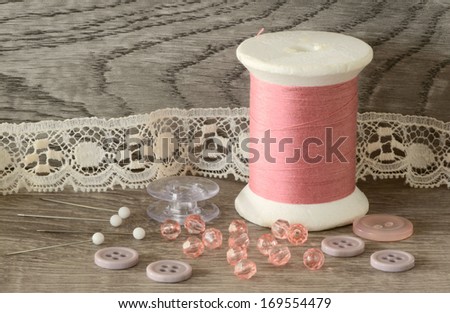 Close up of pink cotton thread spool, beads, and buttons on wooden surface and background
