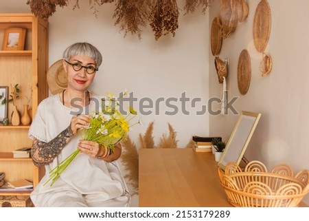 Beautiful retired 60 year old asian woman with short gray hair wearing glasses healthy dazzling clear face charming relaxed lively arranged flowers at home smiling happily looking at the camera. 商業照片 © 