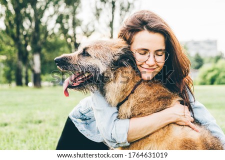 Women hugging dog in the summer park. Cheerful lady with long dark hair in blue jacket hugs and strokes friendly old dog sitting on lush green meadow of public garden on nice day. 