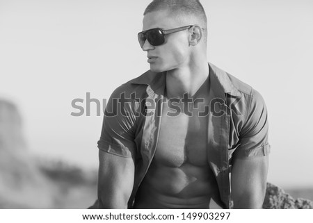 Black and white photo. Young muscular man sitting on the beach. He was wearing a shirt and sunglasses.