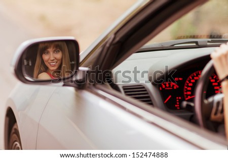 Fashionable girl sitting in a gray car. Her smiling face reflected in the side mirror.