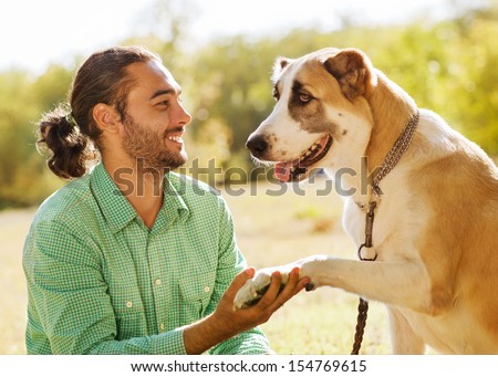 Man and central Asian shepherd in park. He is holding a dog\'s paw and smiles.