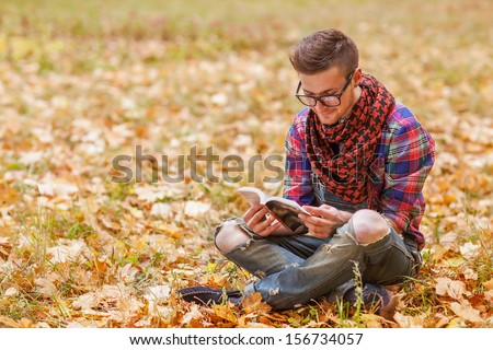 Young hipster relaxed man reading book in nature, back on tree, meadow behind
