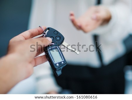 Male hand giving car key to female hand. She is holding a cell phone. In the background, a fragment of the car.