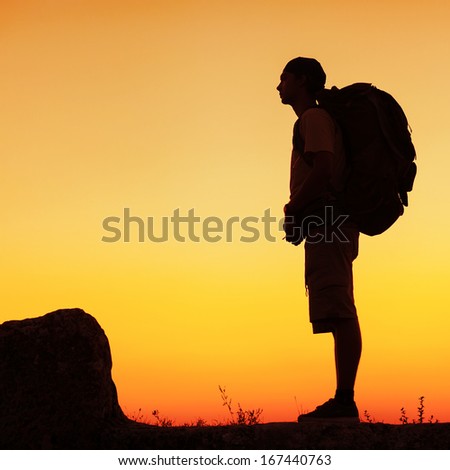 Hiker with backpack standing on top of a mountain and enjoying sunset