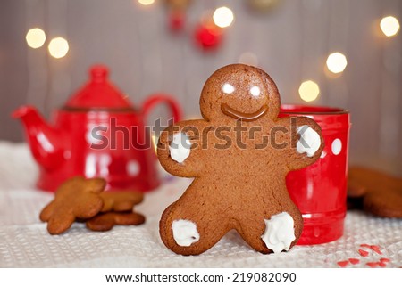 Smiling gingerbread man standing in front of a mug. Teapot and more cookies on the background. Lights and christmas decoration in the background. Red and brown colors