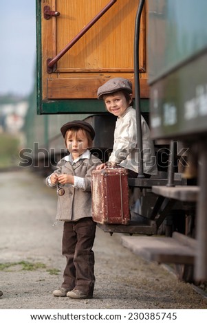 Two boys, dressed in vintage clothing and hat, with suitcase, on a railway station,sitting on steam train stairs