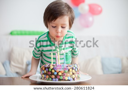 Beautiful adorable four year old boy in green shirt, celebrating his birthday, blowing candles on homemade baked cake, indoor. Birthday party for kids