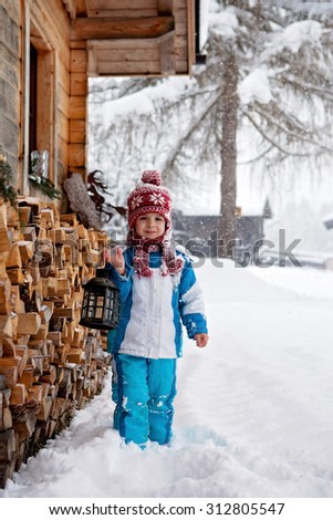 Adorable little boy with lantern, standing in front of a winter cottage while snowing, winter time